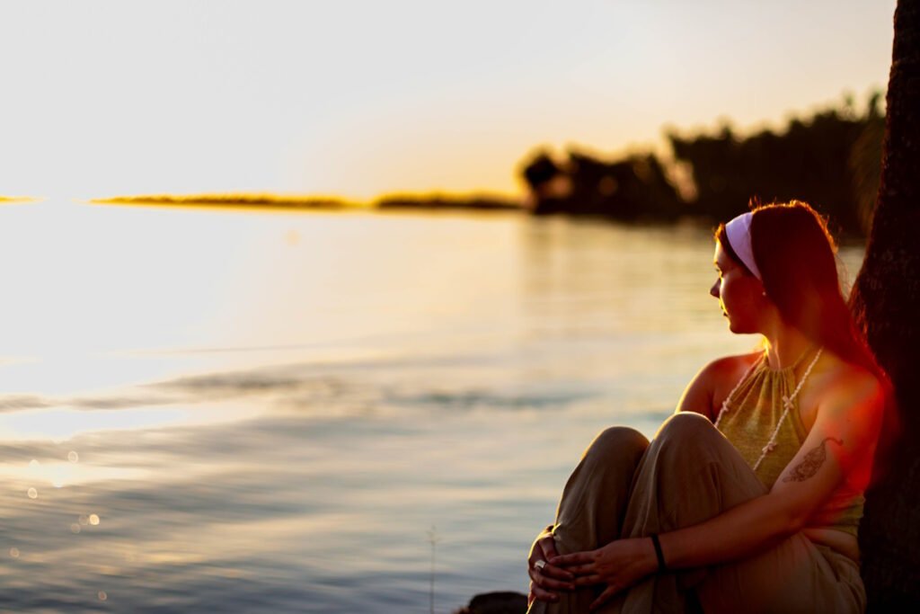 jeune femme assise contre un arbre au bord de l'eau qui regarde le coucher du soleil dans la baie de tapuamu à taha'a en polynésie française