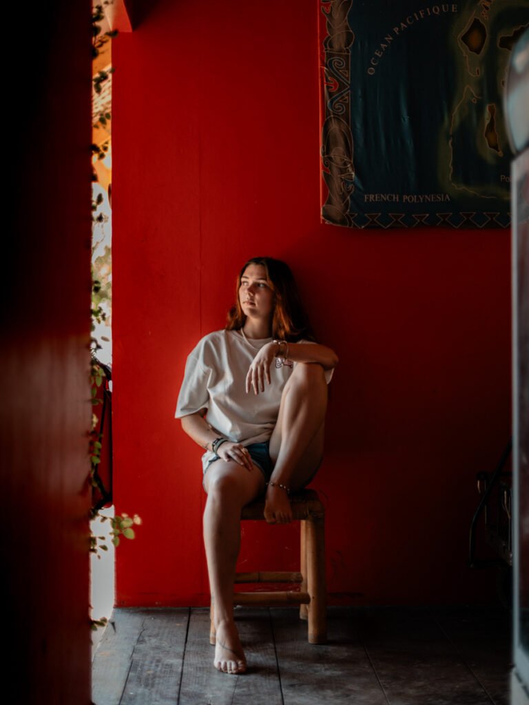 jeune femme aux cheveux long en short assise sur un tabouret qui regarde vers l'extérieur en arrière plan un mur rouge elle est à moitié dans la pénombre