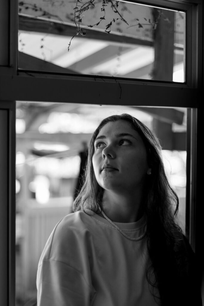 portrait en noir et blanc d'une jeune femme aux cheveux long assise sur le bord d'une fenêtre et regarde vers l'extérieur