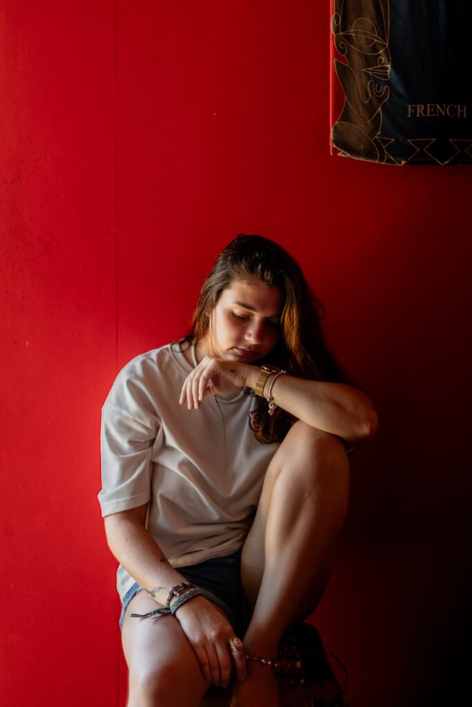 jeune femme en pleine reflexion aux cheveux long en short assise sur un tabouret en arrière plan un mur rouge elle est à moitié dans la pénombre