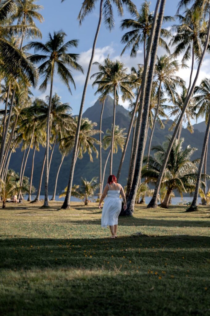 une jeune femme aux cheveux rouge avec une longue robe blanche en train de marcher au milieu des cocotiers sur une plage à moorea en polynésie française en lumière naturelle